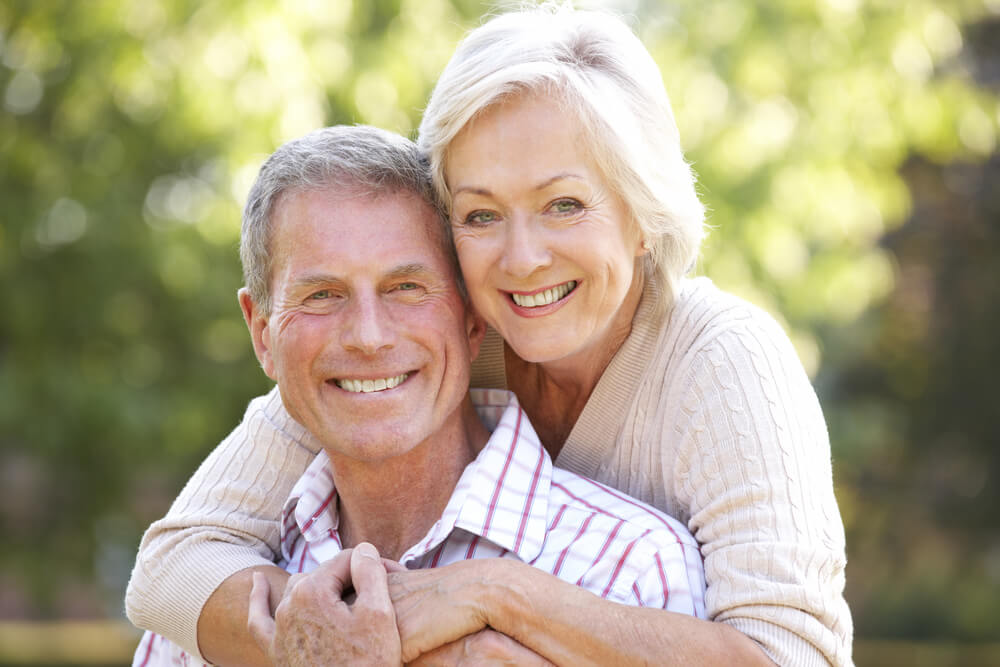 Smiling senior couple outside. Woman is behind the man with her arms wrapped around him.